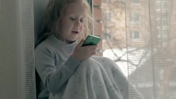 Heureuse petite fille aux cheveux roux ondulés assise sur le rebord de la fenêtre, couvrant une couverture et utilisant le téléphone, parlant, appel vidéo, portrait en gros plan — Video