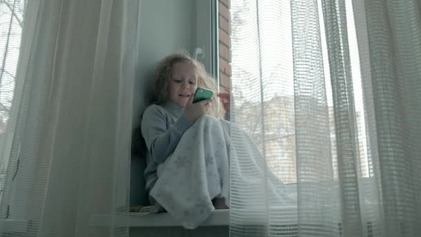 Niña feliz con el pelo rojo ondulado se sienta en el alféizar de la ventana, cubriendo una manta y usando el teléfono, hablando, videollamadas — Vídeos de Stock