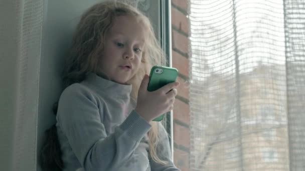 Heureuse petite fille aux cheveux roux ondulés assise sur le rebord de la fenêtre, couvrant une couverture et utilisant le téléphone, parlant, appel vidéo, portrait en gros plan — Video