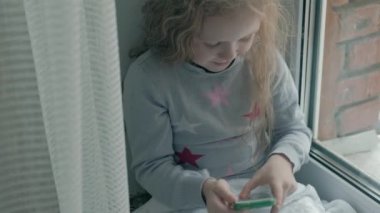 pencere kenarında otururken, bir battaniye kapsayan ve telefonla konuşurken, dalgalı Kızıl saçlı video arama, yakın çekim portre mutlu küçük kız