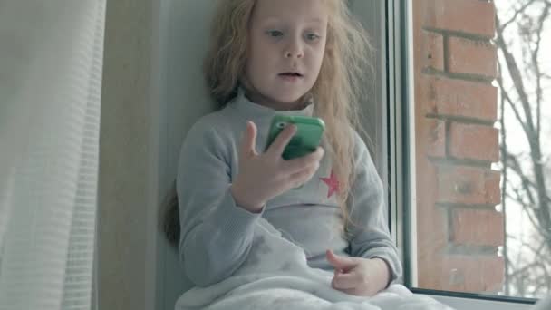Ευτυχισμένο κοριτσάκι με κυματιστή κόκκινα μαλλιά κάθεται στο περβάζι, που καλύπτει μια κουβέρτα και χρησιμοποιώντας το τηλέφωνο, μιλώντας, βίντεο κλήσης, close-up πορτρέτο — Αρχείο Βίντεο