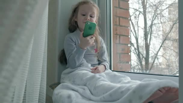 Niña feliz con el pelo rojo ondulado se sienta en el alféizar de la ventana, cubriendo una manta y usando el teléfono, hablando, videollamadas — Vídeos de Stock