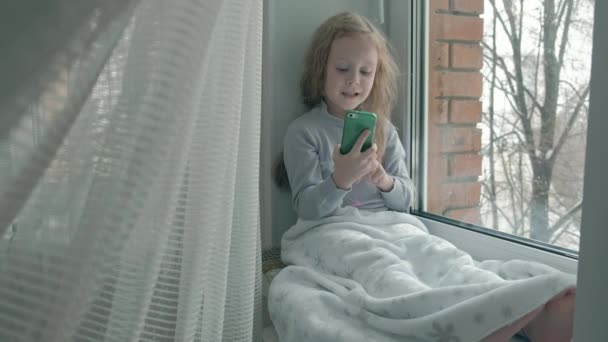Щаслива маленька дівчинка з хвилястим червоним волоссям сидить на підвіконні, покриваючи ковдрою і використовуючи телефон, розмовляючи, відеозв'язуючи — стокове відео