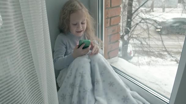 ウェーブのかかった赤い髪を持つ少女は、窓辺に、毛布をカバーし、電話、会話、ビデオ通話を使用して座っている幸せ — ストック動画