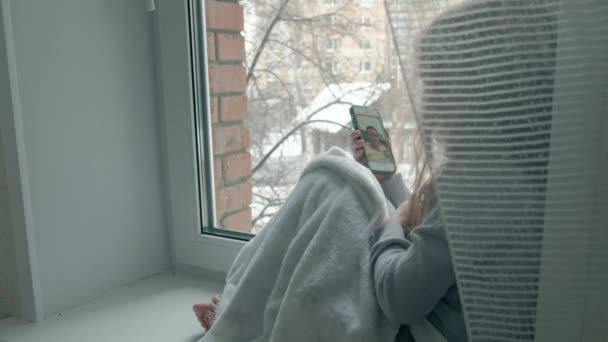 Heureuse petite fille aux cheveux roux ondulés assise sur le rebord de la fenêtre, couvrant une couverture et utilisant le téléphone, parlant, appel vidéo, portrait en gros plan — Video