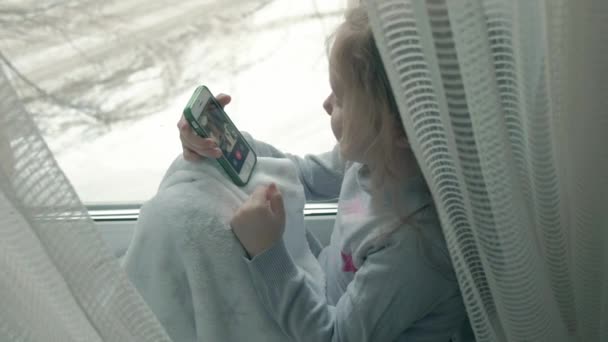 Heureuse petite fille aux cheveux roux ondulés assise sur le rebord de la fenêtre, couvrant une couverture et utilisant le téléphone, parlant, appel vidéo, portrait en gros plan — Video