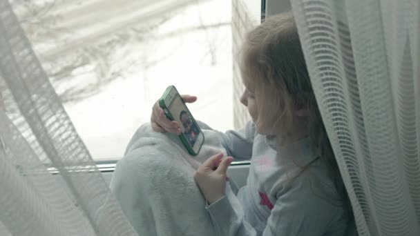 Szczęśliwa dziewczynka z faliste czerwone włosy siedząc na parapecie, obejmujący koc i przez telefon, rozmowa, wideorozmowy, szczegół portret — Wideo stockowe