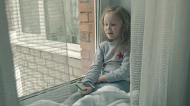 mutlu küçük kız dalgalı Kızıl saçlı bir battaniye kapsayan ve kullanarak telefon, söz, video istek pencere üzerinde oturur