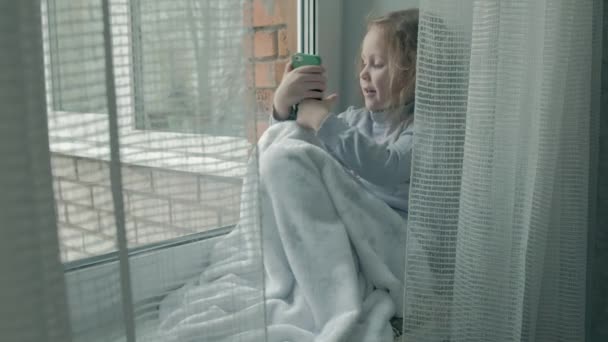 Heureuse petite fille aux cheveux roux ondulés s'assoit sur le rebord de la fenêtre, couvrant une couverture et utilisant le téléphone, parlant, appel vidéo — Video