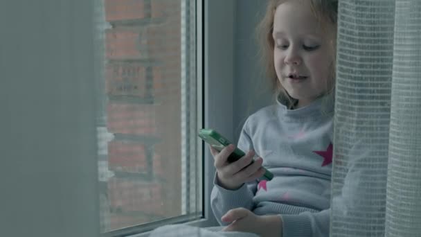 Heureuse petite fille aux cheveux roux ondulés assise sur le rebord de la fenêtre, couvrant une couverture et utilisant le téléphone, parlant, appel vidéo, portrait en gros plan — Video