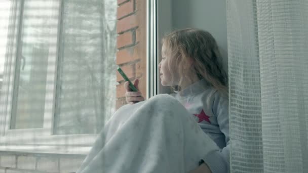 Heureuse petite fille aux cheveux roux ondulés assise sur le rebord de la fenêtre, couvrant une couverture et utilisant le téléphone, parlant, appel vidéo, portrait en gros plan — Video