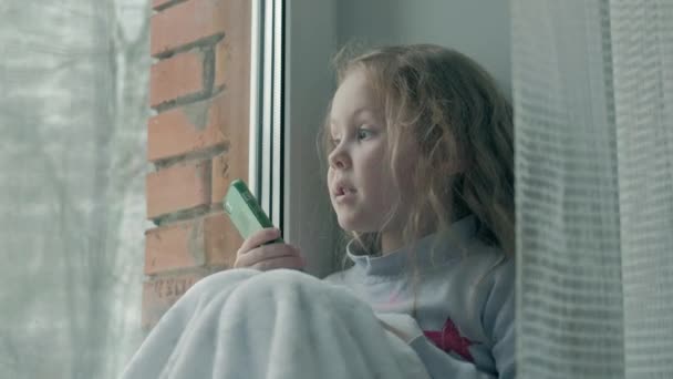Boldog kis lány, hullámos, vörös haja ül az ablakpárkányon, amely egy takarót, és a telefont, beszél, video hívás, közeli portré — Stock videók