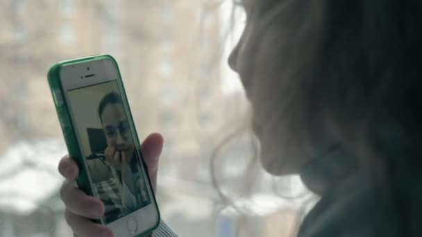 Niña feliz con el pelo rojo ondulado sentado en el alféizar de la ventana, cubriendo una manta y utilizando el teléfono, hablando, videollamadas, primer plano retrato — Vídeos de Stock