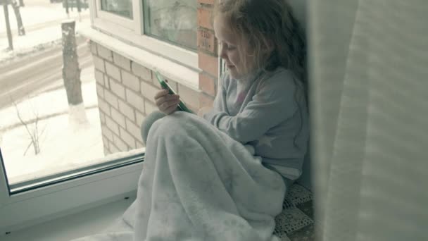 Счастливая маленькая девочка с волнистыми рыжими волосами сидит на подоконнике, прикрывая одеяло и используя телефон, разговаривая, видео-звонки — стоковое видео