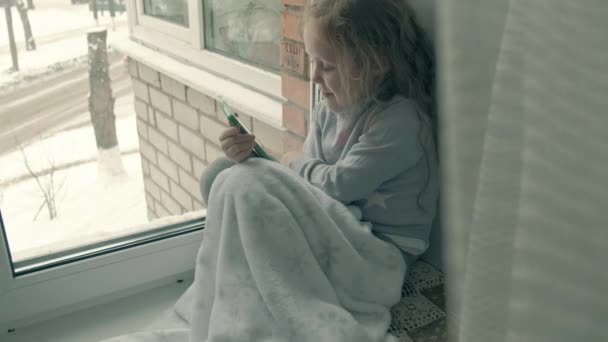 Niña feliz con el pelo rojo ondulado se sienta en el alféizar de la ventana, cubriendo una manta y usando el teléfono, hablando, videollamadas — Vídeo de stock