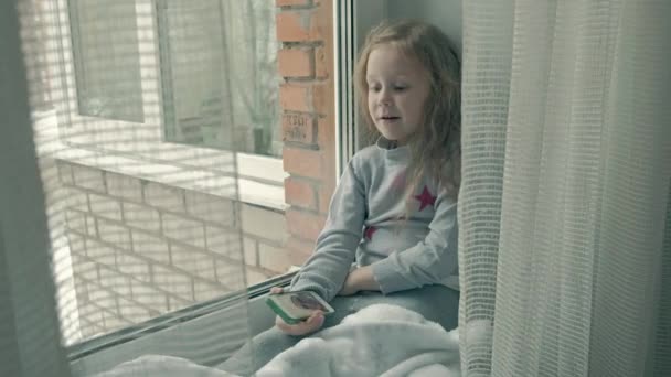 Heureuse petite fille aux cheveux roux ondulés s'assoit sur le rebord de la fenêtre, couvrant une couverture et utilisant le téléphone, parlant, appel vidéo — Video