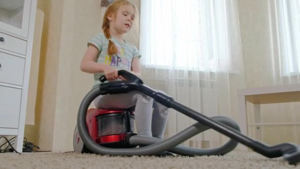En liten flicka med blont hår sitter på en dammsugare och rensar upp, ger ordning och renlighet, hjälper mamma — Stockvideo