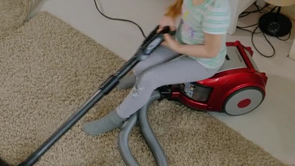 Uma menina com cabelo loiro senta-se em um aspirador de pó e limpa, traz ordem e limpeza, ajuda a mãe — Vídeo de Stock