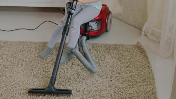 Seorang gadis kecil berambut pirang duduk di atas vacuum cleaner dan membersihkan, membawa ketertiban dan kebersihan, membantu ibu — Stok Video