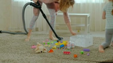 Anne ve kızı, bir elektrik süpürgesi ile genç bir kadın temizler, sarı saçlı küçük bir kız oyuncaklar, bir kap içinde yardımcı olur anne Tasarımcısı toplar.
