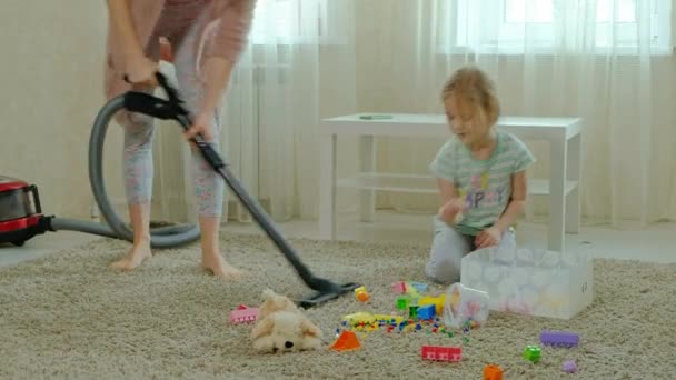 Mamma och dotter, en ung kvinna rensar upp med en dammsugare, en liten flicka med blont hår samlar leksaker, designer i en behållare, hjälper mamma — Stockvideo