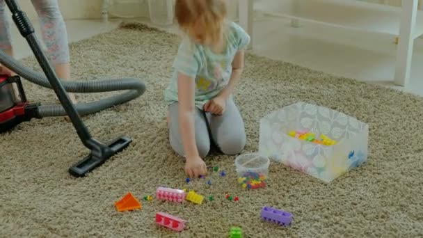Anne ve kızı, bir elektrik süpürgesi ile genç bir kadın temizler, sarı saçlı küçük bir kız oyuncaklar, bir kap içinde yardımcı olur anne Tasarımcısı toplar. — Stok video