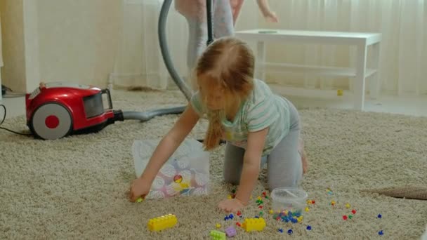 Mãe e filha, uma jovem mulher limpa com um aspirador de pó, uma menina com cabelo loiro coleta brinquedos, o designer em um recipiente, ajuda a mãe — Vídeo de Stock