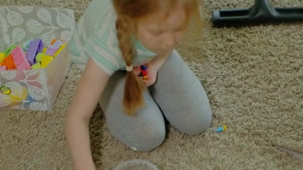 妈妈和女儿, 一个年轻的女人用吸尘器清理, 一个金发碧眼的小女孩收集玩具, 设计师在一个容器里, 帮助妈妈 — 图库视频影像