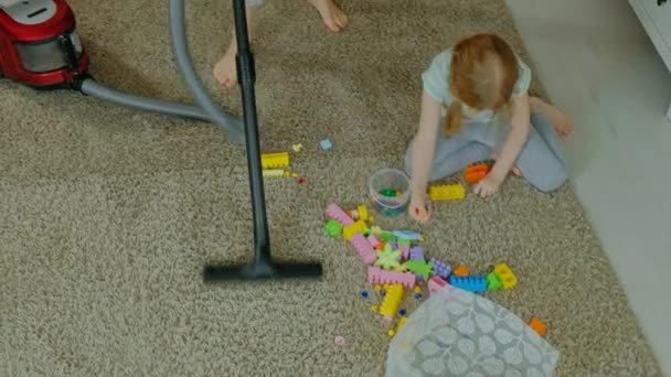 Ibu dan anak, seorang wanita muda membersihkan dengan vacuum cleaner, seorang gadis kecil dengan rambut pirang mengumpulkan mainan, desainer dalam wadah, membantu ibu — Stok Video