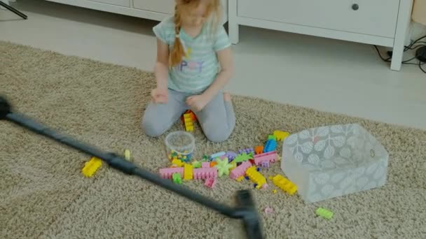 Mamá y su hija, una mujer joven limpia con una aspiradora, una niña con el pelo rubio recoge juguetes, el diseñador en un recipiente, ayuda a mamá — Vídeos de Stock