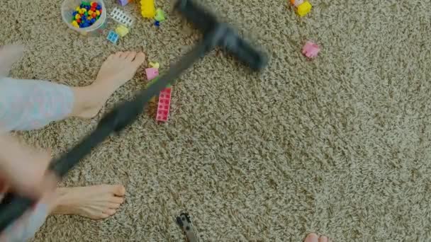 Mamma och dotter, en ung kvinna rensar upp med en dammsugare, en liten flicka med blont hår samlar leksaker, designer i en behållare, hjälper mamma — Stockvideo