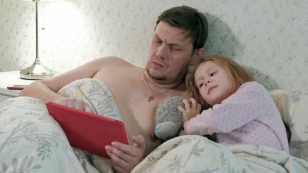 Тато і маленька дочка на ліжку грають на планшеті — стокове відео
