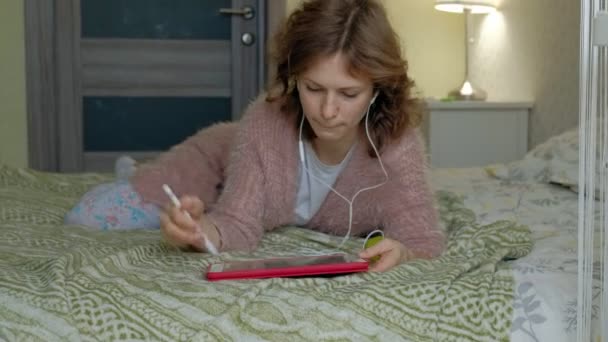 Szczęśliwa dziewczyna słuchanie muzyki i czerpie z rysika na tablecie w sypialni w domu. Tkanina surfing, odległość pracy poczęcia, freelancer — Wideo stockowe