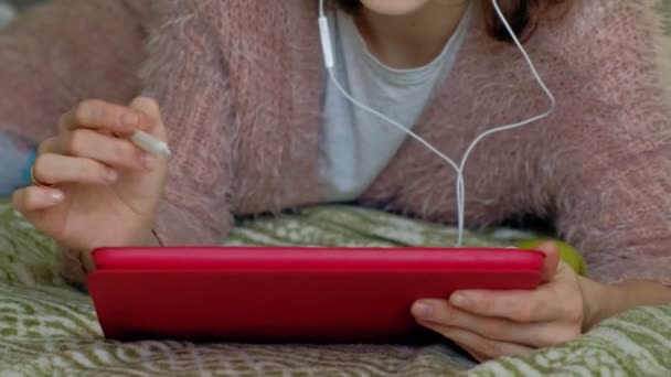 Chica feliz escuchando música y dibuja con un lápiz usando una tableta en el dormitorio en casa. Web surf, concepción del trabajo a distancia, freelancer — Vídeos de Stock