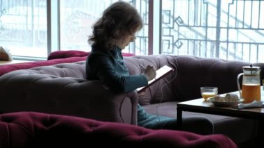 Sarı saçlı, Yeşil elbiseli çekici genç kadın bir tablet kullanarak çalışır, ekran kalemi kullanır, kulaklık ile müzik dinler, bir kafede akşam. Freelancer kavramı, uzak iş.