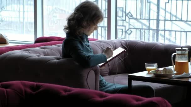 迷人的年轻女子金发 穿着绿色的衣服 工作使用平板电脑 使用手写笔 听音乐与耳机 在咖啡馆用餐 自由职业者的概念 遥远的工作 — 图库视频影像