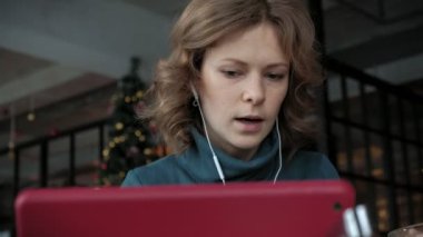 Çalışma, bir kafede yemek yiyor ve kulaklık bir tablet kullanarak ortaklarıyla video konuşmak çekici genç kadın Yeşil elbiseli, sarı saçları ile arıyorum. Freelancer kavramı, uzak iş. Kapat