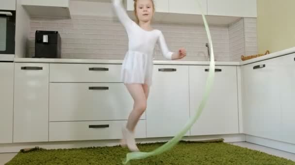 En glad liten tjej i en vit gymnastiska baddräkt tåg, danser med ett band för rytmisk gymnastik, hopp och utför professionell övningar. — Stockvideo