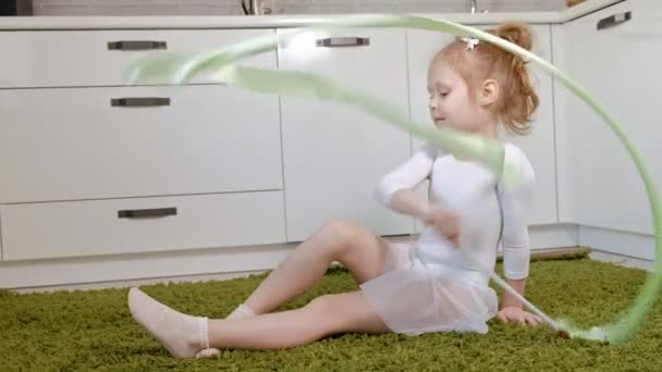 A happy little girl in a white gymnastic swimsuit trains, dances with a ribbon for rhythmic gymnastics, jumps and performing professional exercises. — Stock Video