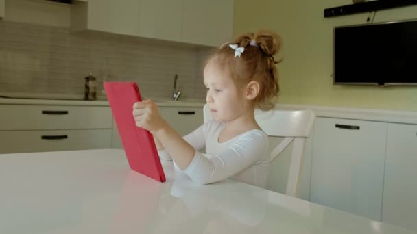 Cute little girl sitting in the kitchen or room at the table and playing using a tablet computer. Web surfing. — Stock Video