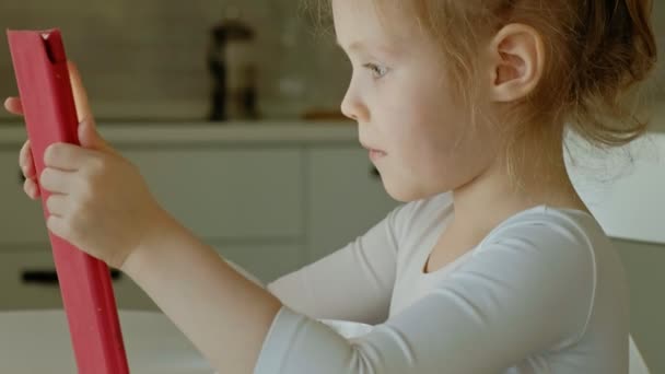 Söt liten flicka sitter i köket eller rum vid bordet och spela med en Tablet PC-dator. Webbsurfning. — Stockvideo