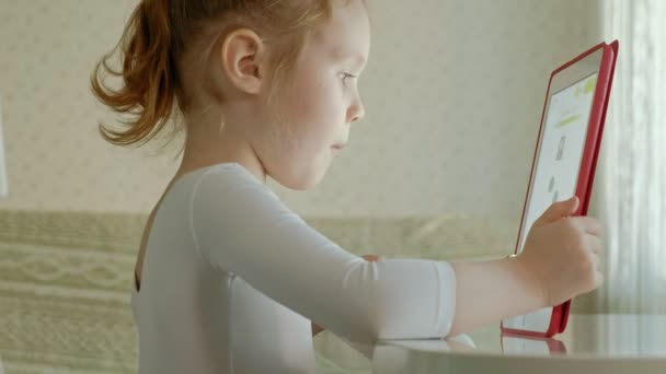 Söt liten flicka sitter i köket eller rum vid bordet och spela med en Tablet PC-dator. Webbsurfning. — Stockvideo