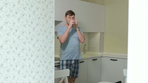 Un hombre en pijama bebiendo agua en la cocina. Buenos días. — Vídeos de Stock