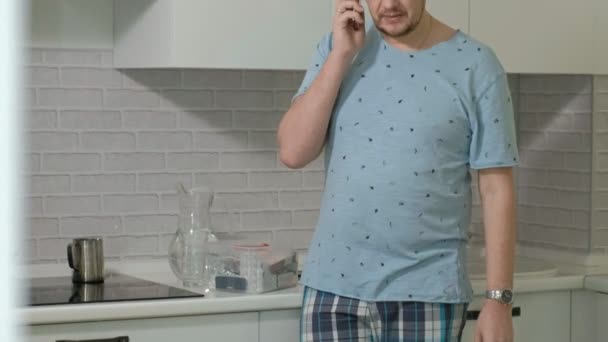 Un hombre en pijama bebiendo agua en la cocina. Buenos días. — Vídeos de Stock