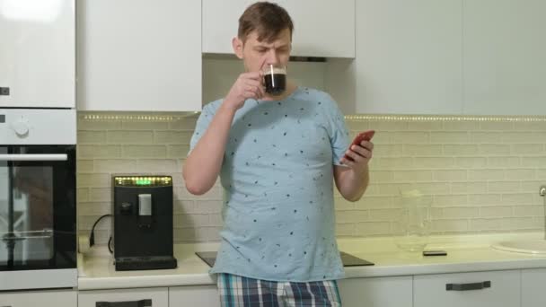 Un hombre en pijama bebiendo agua en la cocina. Buenos días. — Vídeo de stock