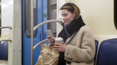 Genç esmer kadın toplu taşıma araçlarına biniyor, kulaklıkla telefonu kullanıyor.