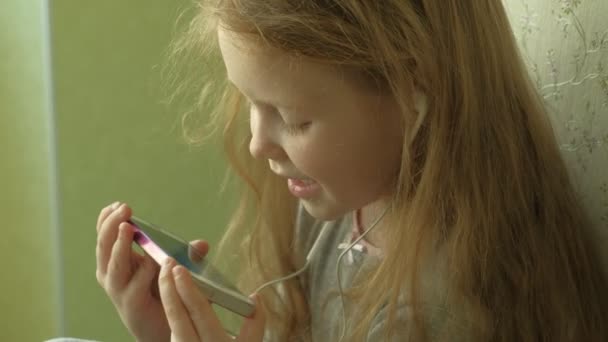 Μικρό κορίτσι κάθεται στο παράθυρο χρησιμοποιεί το τηλέφωνο — Αρχείο Βίντεο