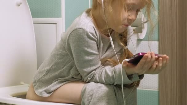 Klein meisje zit op het toilet en maakt gebruik van de telefoon — Stockvideo