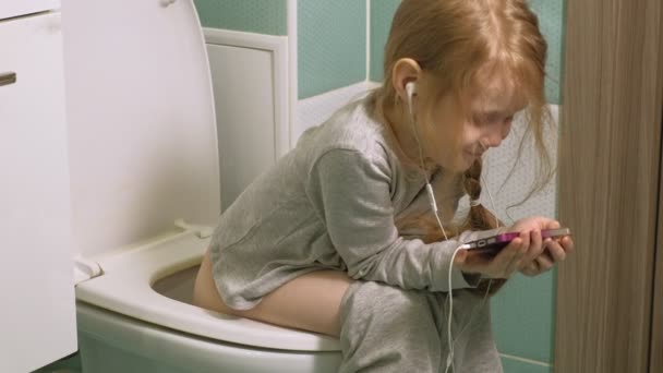 Niña se sienta en el inodoro y usa el teléfono — Vídeos de Stock