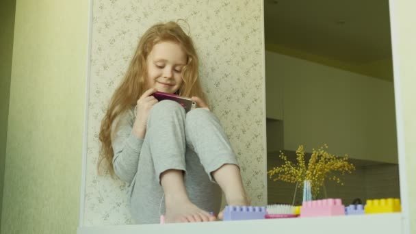 Little girl sitting on the window uses the phone — Stock Video
